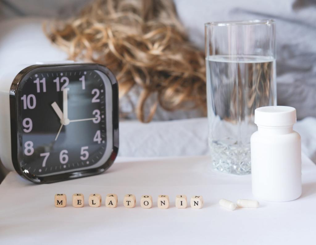 Bedside with a clock, some melatonin capsules, and the word melatonin spelled out on dice blocks.