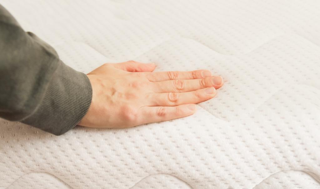 Hand touching and pressing orthopedic mattress on bed.